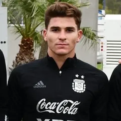Julián Álvarez modelling the Argentina 2026 World Cup home jersey by Adidas — the sky-blue and white vertical stripes carry a three-tone fade that references the nation's three World Cup titles in 1978, 1986 and 2022, with the AFA crest centre-chest and the Adidas Performance logo on the right; the jersey released globally on November 6, 2025 and will be worn at the title defence opener vs Algeria at Arrowhead Stadium in Kansas City on Tuesday June 16, 2026